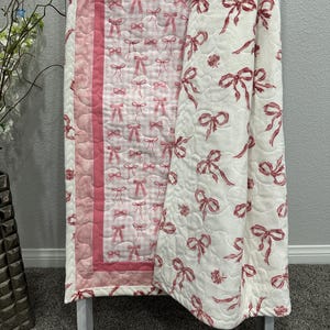 May include: A baby quilt with a pink and white bow pattern. The quilt is displayed on a white wooden ladder. The quilt has a pink border and a gingham pattern on one side. The other side has a repeating pattern of pink bows.