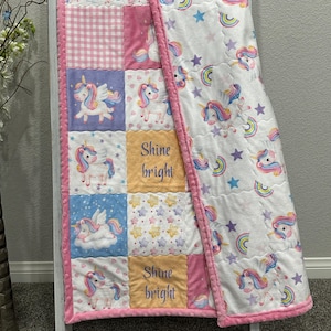May include: A patchwork baby quilt featuring unicorn designs, rainbows, and stars. The quilt has a pink border and backing, with squares of pink, blue, and yellow. The words "Shine bright" are embroidered on two of the squares. The quilt is displayed on a white ladder.