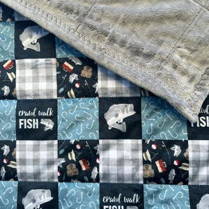May include: A patchwork quilt with a gray fleece backing. The quilt features a blue and white checkered pattern, a black background with fishing lures and fish, and the text "crawl walk FISH".