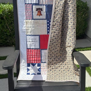 May include: A patriotic quilt draped over a black rocking chair. The quilt features red, white, and blue squares with patriotic symbols and text, including the Liberty Bell and the phrase "We the People." The reverse side has a floral pattern.