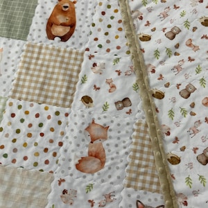 May include: A patchwork quilt featuring woodland animal designs. The quilt showcases a bear, fox, and raccoon, along with other forest elements. The quilt has a variety of patterns, including polka dots and gingham, in shades of brown, beige, and green.
