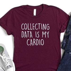 May include: Burgundy t-shirt with white text that reads "Collecting data is my cardio". The shirt is laid flat on a white background with a pair of gray sneakers and a pair of blue jeans.