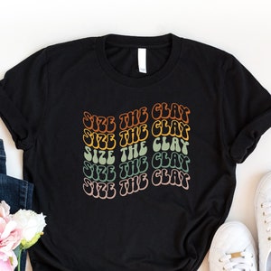 May include: Black t-shirt with the text "Size the Clay" repeated five times in a rainbow gradient. The text is in a retro font style.