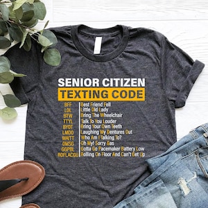May include: A dark gray t-shirt with the text "Senior Citizen Texting Code" in yellow. The text is a list of abbreviations and their meanings, such as BFF, LOL, BTW, TTYL, BYOT, LMDO, WAITT, OMSG, GGPBL, and ROFLACGU.