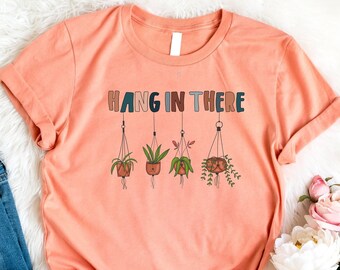 May include: A peach colored t-shirt with the text "HANG IN THERE" in colorful letters. The text is surrounded by a graphic of five hanging plants.