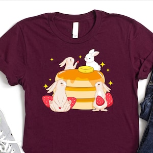 K&ouml;nnte beinhalten: Ein maroonfarbenes T-Shirt mit einem Motiv von drei wei&szlig;en Kaninchen, die einen Stapel Pfannkuchen mit Butter und Erdbeeren essen.