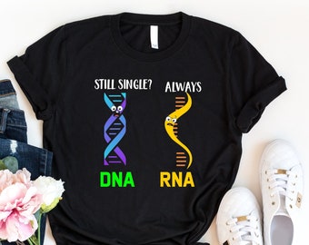 以下が含まれることがあります： DNAの鎖が2本描かれた黒いTシャツ。1本は「DNA」とラベル付けされ、もう1本は「RNA」とラベル付けされている。DNAの鎖には顔があり、その上に「まだ独身？」という文字が書かれている。RNAの鎖には顔があり、その上に「いつも」という文字が書かれている。