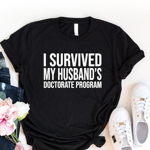 May include: Black t-shirt with white text that reads "I survived my husband's doctorate program". The shirt is laid flat on a white surface with a pair of white sneakers, a bouquet of pink flowers, and a pair of blue jeans.