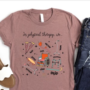 May include: A mauve t-shirt with the text "In physical therapy, we..." and illustrations of physical therapy equipment. The shirt is paired with blue jeans and white sandals.
