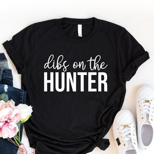 May include: Black t-shirt with white text that reads "dibs on the HUNTER". The shirt is laid flat on a white surface with a pair of white sneakers, blue jeans, and pink flowers.