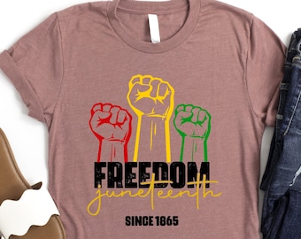 May include: A mauve t-shirt with a graphic of three raised fists in red, yellow, and green. The text "FREEDOM Juneteenth SINCE 1865" is printed below the fists.
