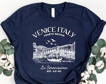 May include: A navy blue t-shirt with a white graphic design of the Rialto Bridge in Venice, Italy. The text "Venice Italy Veneto Region La Serenissima Est. 421 AD" is printed on the shirt.