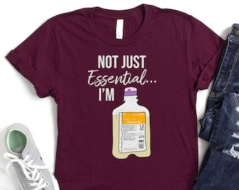 Op de afbeelding: Bordeauxkleurig T-shirt met de tekst "Not Just Essential... I'm" en een cartoonafbeelding van een infuuszak.