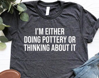 May include: Dark gray t-shirt with the text "I'M EITHER DOING POTTERY OR THINKING ABOUT IT" in white. The shirt is paired with blue jeans and black sandals with brown soles. Greenery is in the background.