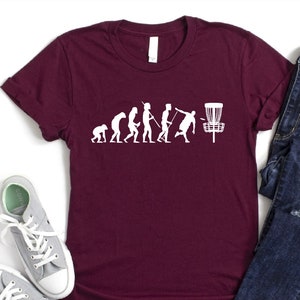 May include: A maroon t-shirt with a white graphic of the evolution of man ending with a person throwing a frisbee. The graphic includes a disc golf basket.