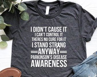 May include: A dark gray t-shirt with white text that reads "I didn't cause it I can't control it There's no cure for it I stand strong =ANYWAY= Parkinson's Disease AWARENESS". The shirt is folded and laid on a wooden surface with a pair of brown sandals and blue jeans.