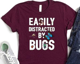 May include: Burgundy t-shirt with white text that reads "Easily Distracted by Bugs". The shirt features a variety of colorful bugs, including a butterfly, a ladybug, and a spider.