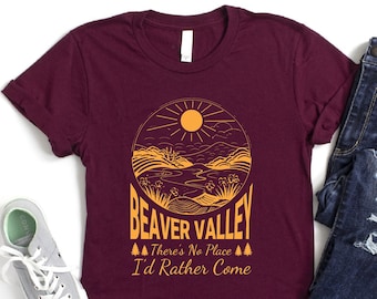 May include: A maroon t-shirt with a graphic design of a sun, mountains, and a river. The text "Beaver Valley" is written in a yellow font. Below the graphic, the text "There's No Place I'd Rather Come" is written in a smaller yellow font.