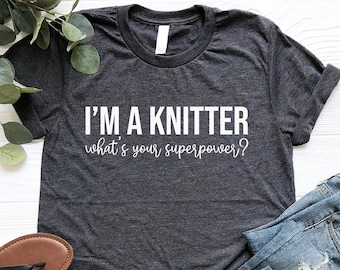 May include: A dark gray t-shirt with the text "I'm a knitter what's your superpower?" in white. The shirt is laying on a white wooden surface with a pair of black sandals and blue jeans.