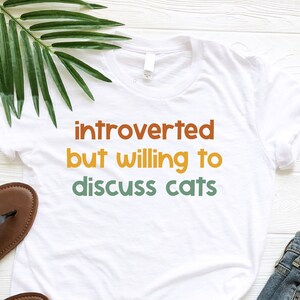 May include: White t-shirt with the text "introverted but willing to discuss cats" in a retro font with orange, yellow, and green colors.