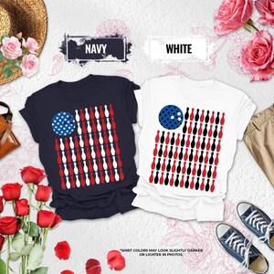 May include: Two short-sleeved t-shirts, one navy and one white, each featuring a graphic design of a bowling ball and bowling pins arranged to resemble an American flag. The navy shirt has white and red pins, while the white shirt has red and black pins.