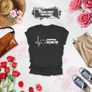 May include: A dark grey heather t-shirt featuring a white graphic of a heartbeat line connected to a heart shape and a school bus. The text DARK GREY HEATHER is printed in a black rectangular box above the shirt, which is surrounded by roses.