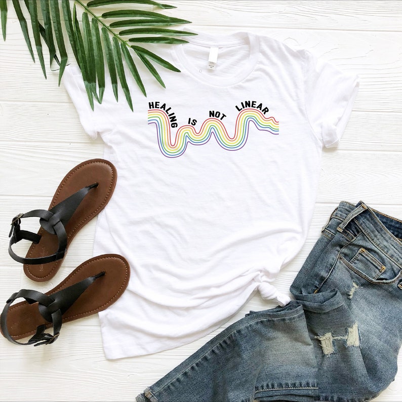 Op de afbeelding: Wit T-shirt met een regenbooggrafiek en de tekst "Healing is not linear".