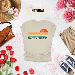 May include: A beige t-shirt with a retro design featuring a sunset, horizontal stripes, and the text "MASTER BUILDER." The image includes a straw hat, a brown leather bag, khaki pants, and navy blue sneakers.