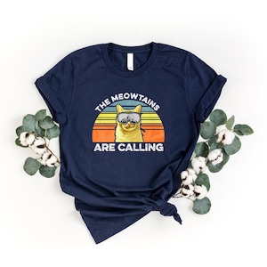 May include: A navy blue t-shirt with a retro sunset graphic featuring a cat wearing ski goggles and the text "The Meowtains Are Calling".