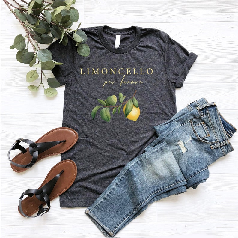 May include: A dark heather grey t-shirt with the text "Limoncello per favore" in gold lettering. A small lemon branch with a yellow lemon is printed on the shirt.