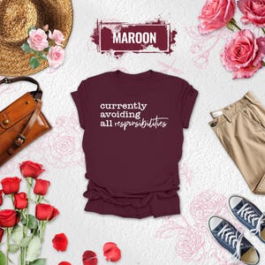 May include: Maroon t-shirt with the text "currently avoiding all responsibilities" in white script. The shirt is styled with khaki pants, blue sneakers, a brown leather bag, a straw hat, and roses.