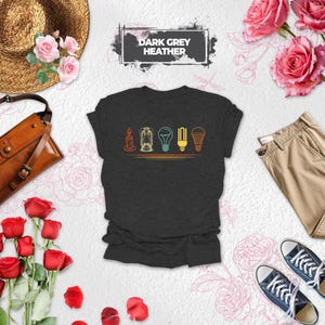 May include: A dark grey heather t-shirt featuring a graphic print of five different light sources, including a candle, a lantern, and three types of light bulbs, arranged in a horizontal row above two thin orange lines with the text DARK GREY HEATHER.