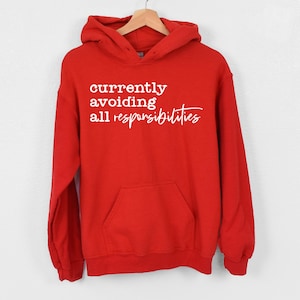 May include: A bright red hooded sweatshirt featuring a front kangaroo pocket and long sleeves. The chest area displays white text that reads currently avoiding all responsibilities in a combination of block and cursive lettering styles.