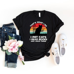 May include: Black t-shirt with a retro sunset graphic featuring a cat reading a book. The text on the shirt reads "That's what I do I pet cats I read books and I know things."