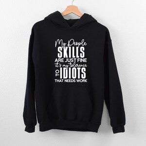 May include: A black hooded sweatshirt featuring white text on the front that reads: My People SKILLS ARE JUST FINE it's my tolerance TO IDIOTS THAT NEEDS WORK. The garment includes a front kangaroo pocket, long sleeves, and a drawstring hood.