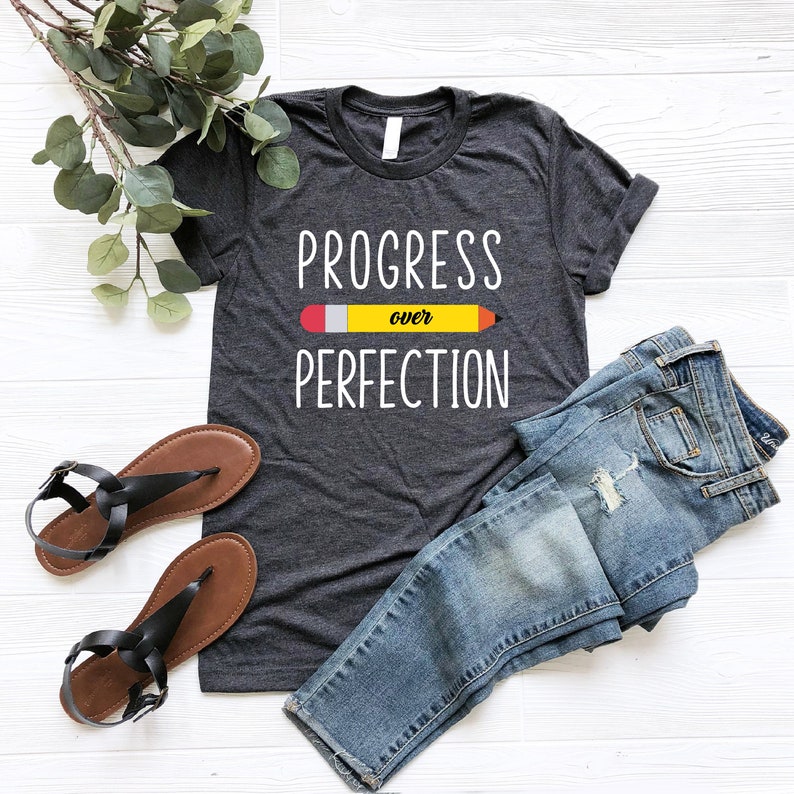 May include: A dark gray t-shirt with the text "PROGRESS over PERFECTION" in white lettering. A yellow pencil with a red eraser is drawn above the word "over".