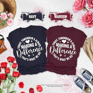 May include: Two short-sleeved t-shirts in navy and maroon, each featuring white text that reads "TODAY, TOMORROW & ALWAYS MAKING A DIFFERENCE THAT'S WHAT WE DO" with a heart and star accents. The shirts are displayed alongside a straw hat, a brown bag, khaki pants, blue sneakers, and pink roses.