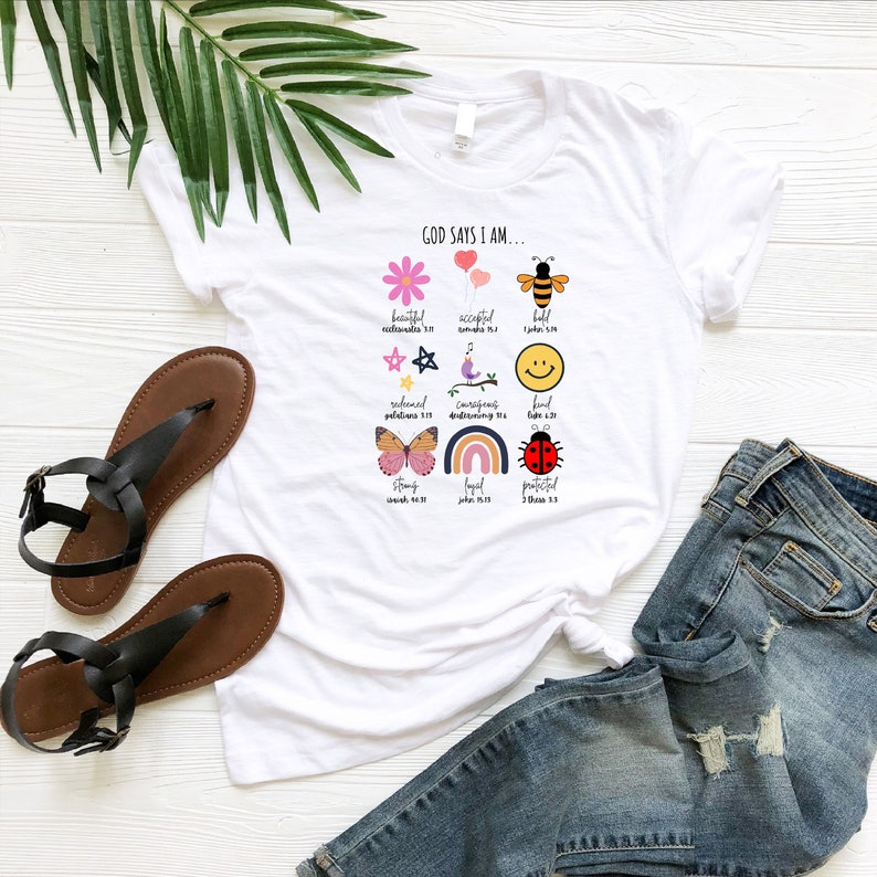 K&ouml;nnte beinhalten: Wei&szlig;es T-Shirt mit dem Text "Gott sagt, ich bin..." und bunten Illustrationen einer Blume, Ballons, einer Biene, eines Sterns, eines Vogels, eines Smiley-Gesichts, eines Schmetterlings, eines Regenbogens und eines Marienk&auml;fers. Zu jeder Illustration geh&ouml;rt ein passender Bibelvers.