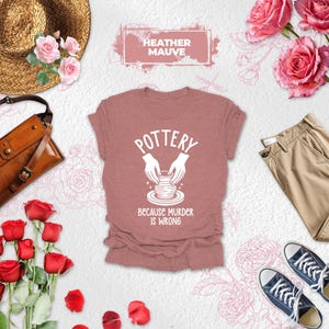 May include: Heather mauve t-shirt with the text "POTTERY" and a graphic of hands shaping clay. The text "BECAUSE MURDER IS WRONG" is below the graphic. The image also includes a straw hat, a brown leather bag, red roses, pink roses, khaki pants, and blue sneakers.