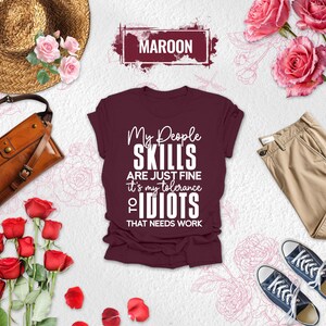 May include: Maroon t-shirt with the text "My People Skills Are Just Fine It's My Tolerance of Idiots That Needs Work." The shirt is displayed with a straw hat, brown leather bag, khaki pants, and navy blue sneakers.