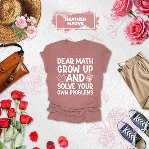 May include: A heather mauve t-shirt with white text that reads Dear Math Grow Up And Solve Your Own Problems, featuring a small graphic of a baseball and a calculator. The shirt is surrounded by a straw hat, a brown leather bag, khaki pants, sneakers, and pink roses.