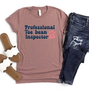 May include: A mauve t-shirt with the text "Professional Toe bean Inspector" in navy blue with a pink paw print.