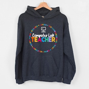 May include: A dark charcoal gray hooded sweatshirt featuring a graphic on the chest. The design includes a white computer icon above the text "Computer Lab TEACHER" in colorful letters, surrounded by a circular border of words like LOVING, FUN, COMPASSIONATE, LOYAL, WARM, DEDICATED, CARING, KIND, and RELIABLE.