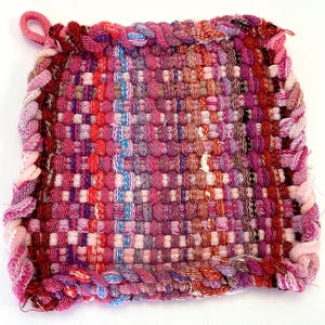 Peut inclure: Un dessous de plat tissé rose et violet avec un bord tressé. Le dessous de plat est fait de fils de différentes couleurs.