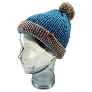 May include: A blue and gray knit beanie with a pom pom on top. The beanie has a brown leather tag with a logo on it.