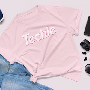 May include: A light pink t-shirt with the word "Techie" in white, outlined in pink. The shirt is styled with blue jeans, navy blue sneakers, a black smartphone, wireless earbuds, and sunglasses.