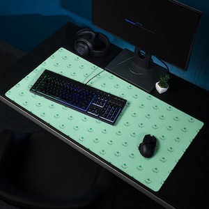 May include: A light green rectangular desk mat with a repeating pattern of small green clover shapes. A black keyboard with illuminated keys, a black mouse, and a computer monitor are on the mat. A pair of black headphones sits nearby.