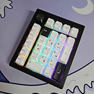 May include: A black and white keyboard with a black frame and a silver knob. The keys are white with black lettering and have a rainbow backlight. The keys are labelled with numbers, letters, and symbols. The keyboard has a row of keys labelled "M1" through "M5".