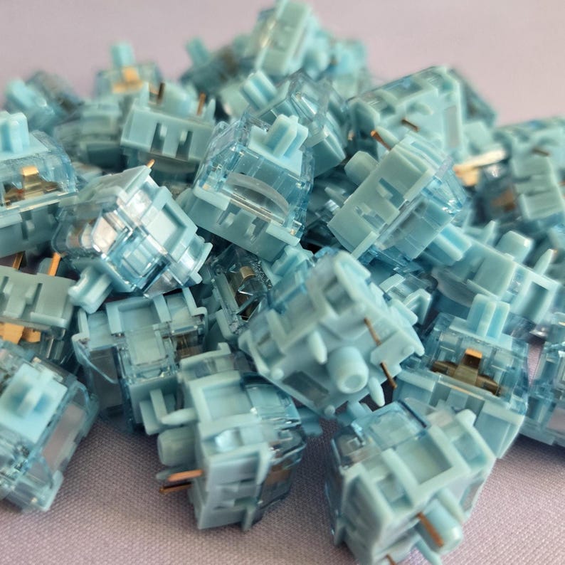 May include: A collection of light blue mechanical keyboard switches featuring translucent plastic housings and visible metal contact pins. Each switch has a central stem and a square base design, creating a uniform pile of components.