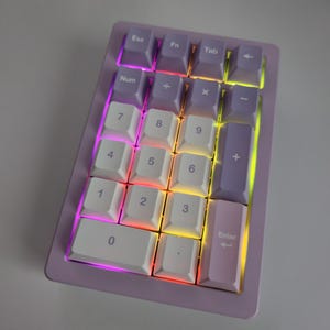 May include: A mechanical number pad featuring a light purple frame and a mix of white and light purple keycaps. The keys are labeled with numbers, mathematical symbols, and function commands. The device emits a vibrant, multi-colored RGB backlight.